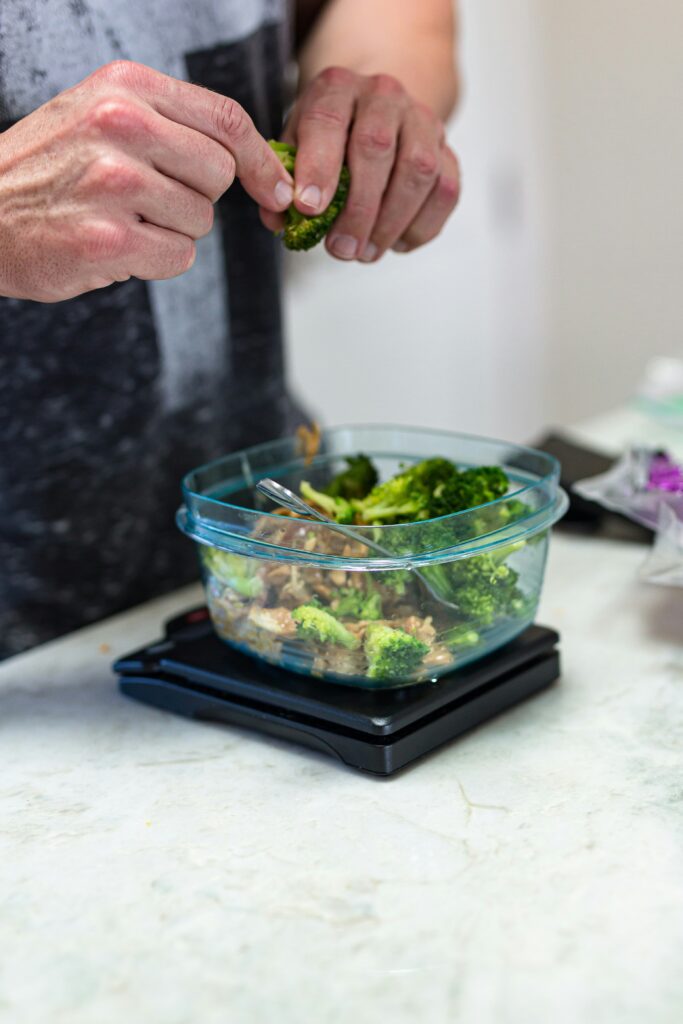 Hand adding broccoli to a glass meal prep container on a kitchen scale during portioning and meal prep