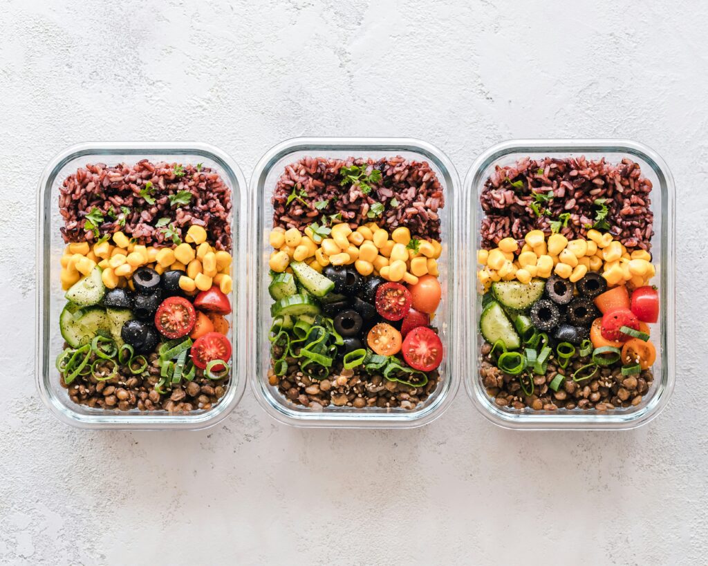 Three glass meal prep containers filled with rice, lentils, corn, olives, cucumbers, tomatoes, and green onions arranged neatly from above