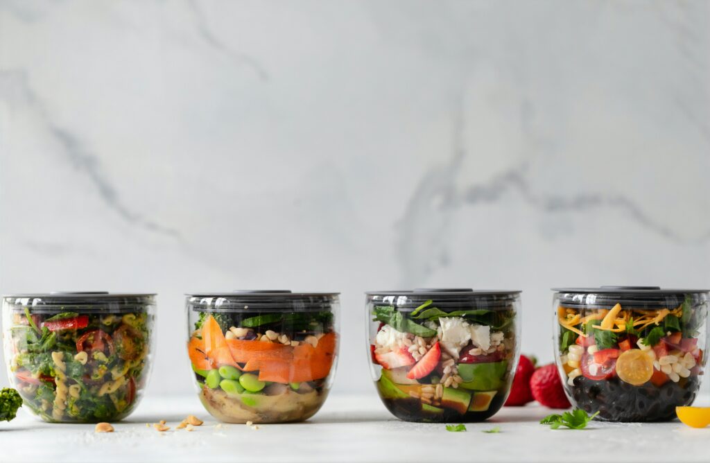 Four glass meal prep jars filled with layered salads, vegetables, grains, beans, and fresh toppings on a clean white surface.
