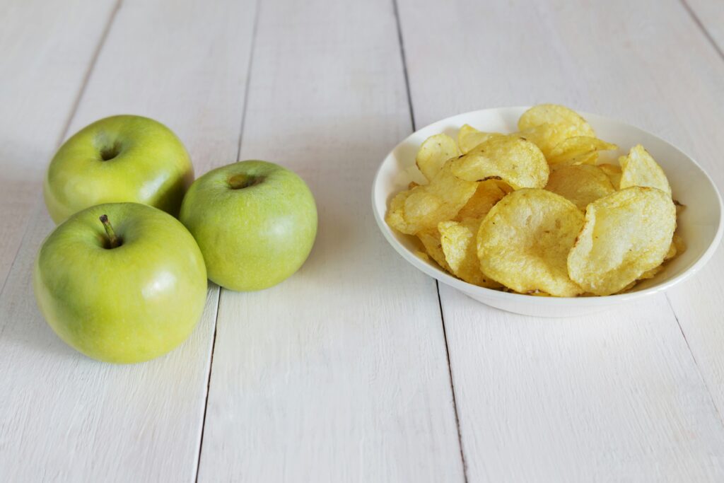 Green apples next to a bowl of potato chips on a light wooden surface, showing a simple healthy snack versus processed snack comparison.